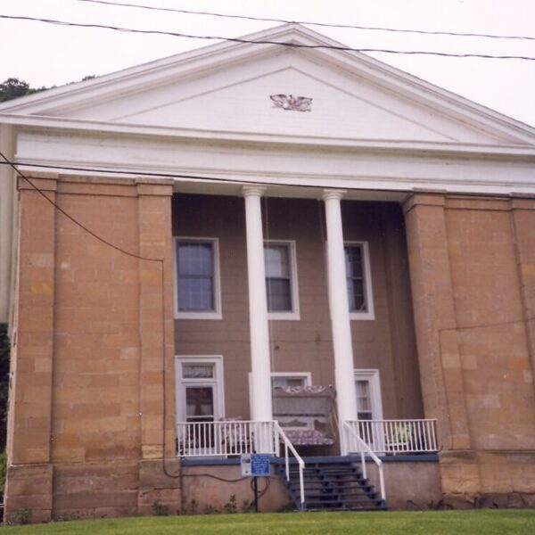 A historic, two-story stone building with four white columns and a triangular pediment. There are steps leading to the entrance, and power lines are visible in front.