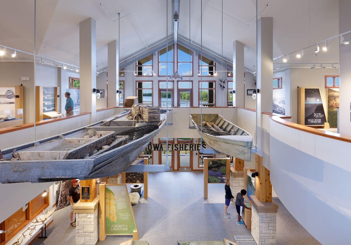 Interior of a modern visitor center with high ceilings, showcasing two suspended boats. Information displays and people are visible on both levels.