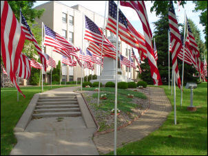 Multiple American flags displayed outdoors in front of a building with steps and a pathway, surrounded by green grass and trees.