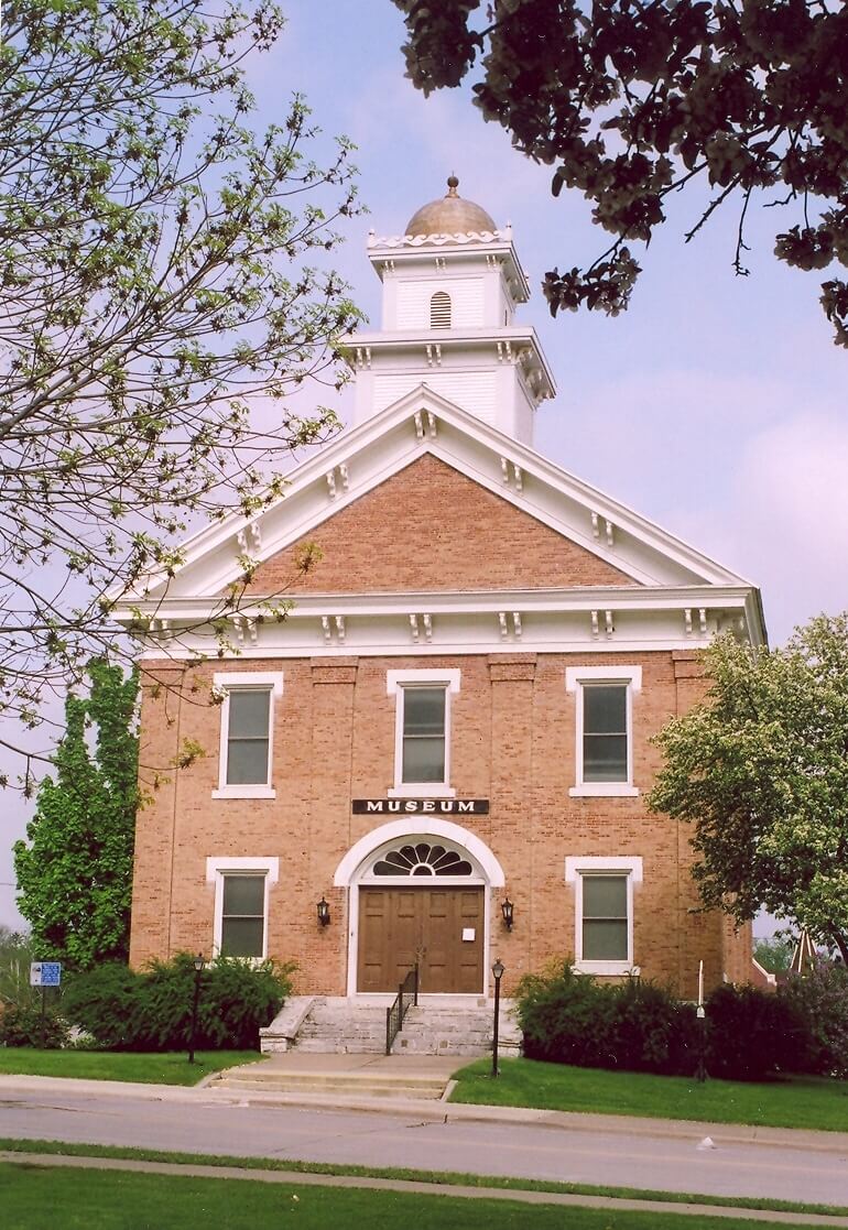 A historic brick building with white trim and a domed belfry, labeled "Museum," surrounded by trees and shrubs.