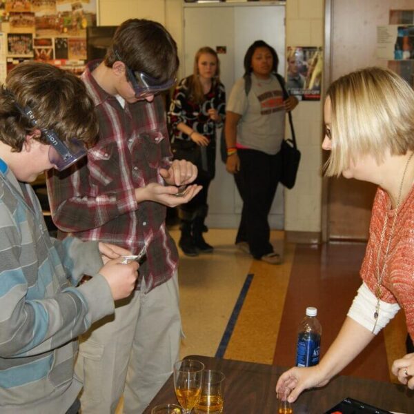 Students wearing safety goggles engage in a science experiment with a teacher in a classroom. Other students are visible in the background.