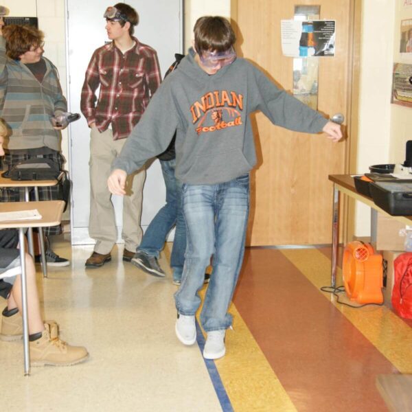 A person wearing goggles walks along a taped line on the floor in a classroom, observed by others.