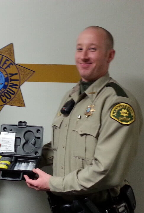 A uniformed officer smiling while holding an open case with a breathalyzer inside in front of a wall with a sheriff's department logo.