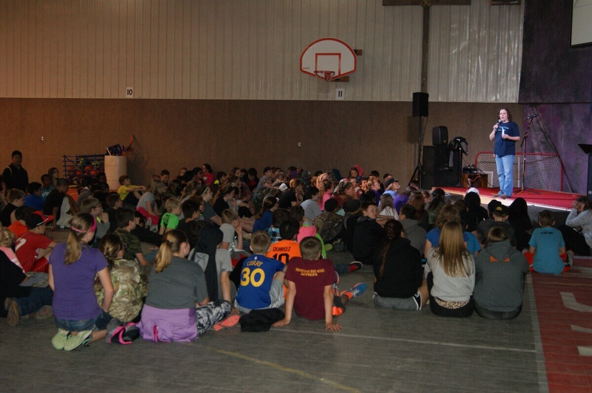 A large group of children sits on a gym floor facing a speaker on stage holding a microphone. A basketball hoop is visible in the background.