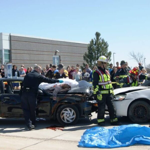 Emergency responders attend to a simulated car accident with two damaged vehicles and a crowd observing, in front of a brick building.