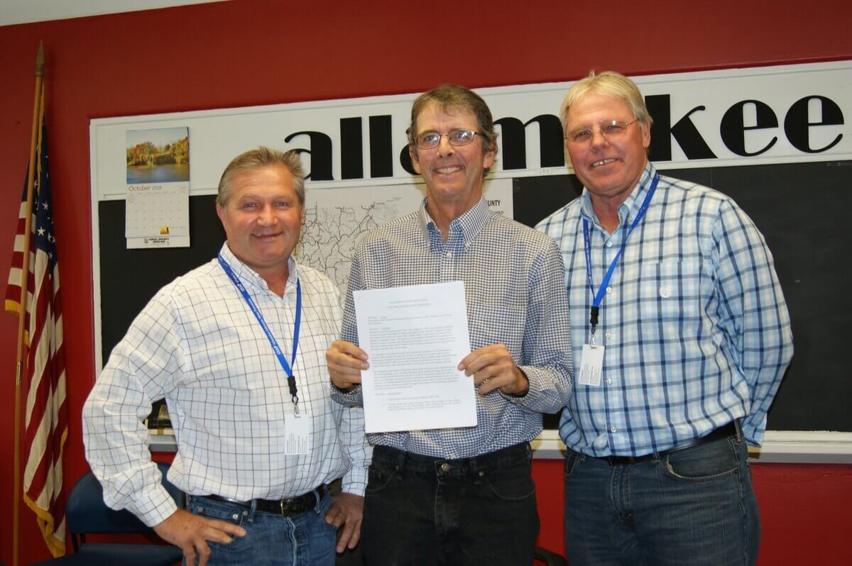 Three men standing indoors, with one holding a document. They're wearing plaid shirts and lanyards. A partial sign and an American flag are visible in the background.