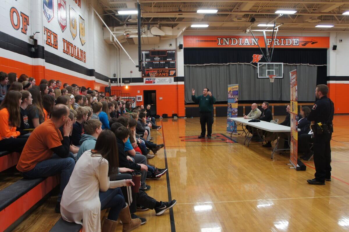 A person speaks to a large group of seated students in a school gymnasium. A panel of four people sits at a table near the speaker. Police officer stands beside the table.