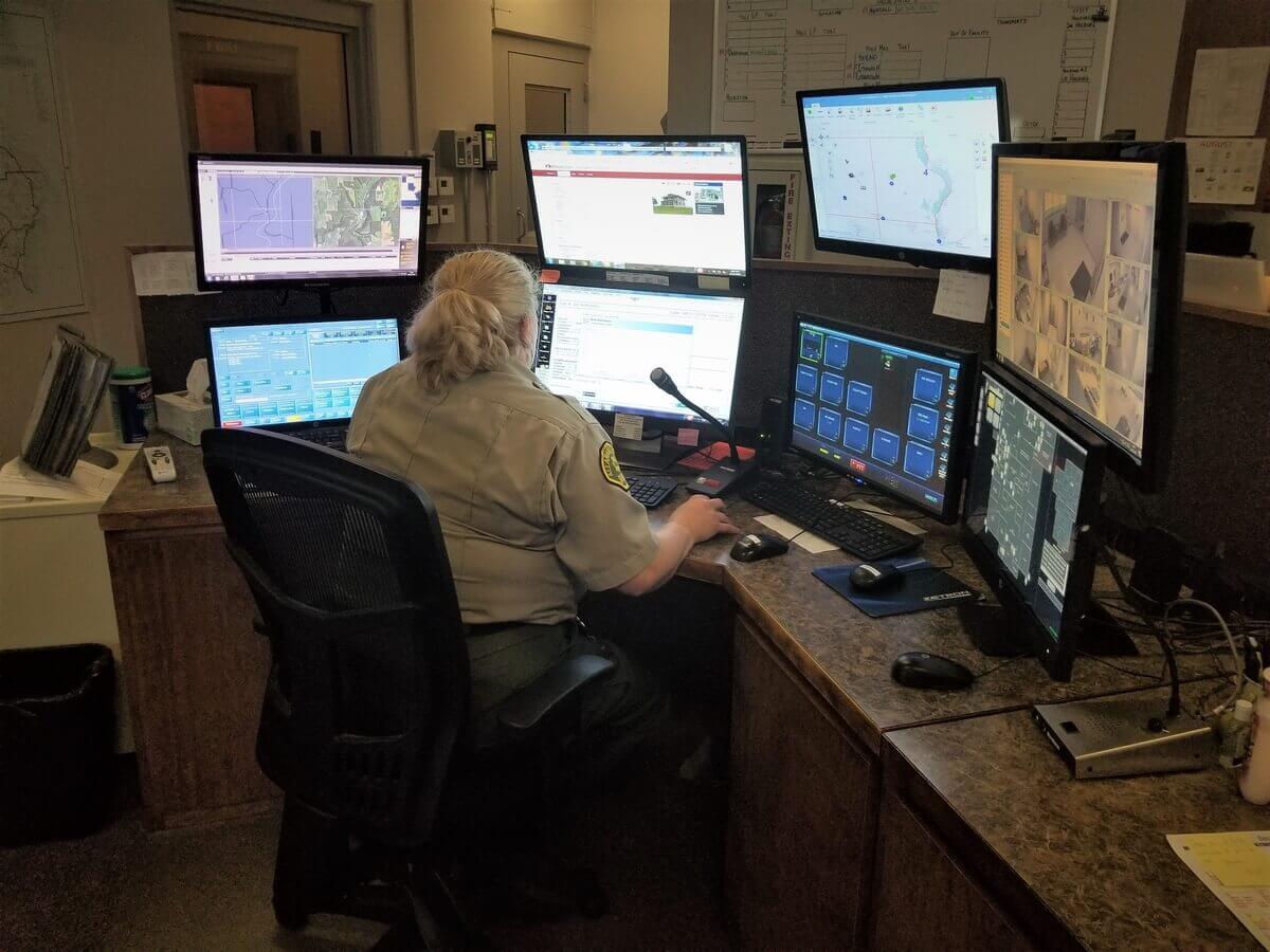 Person in a control room sitting at a desk with multiple monitors displaying maps and surveillance feeds.