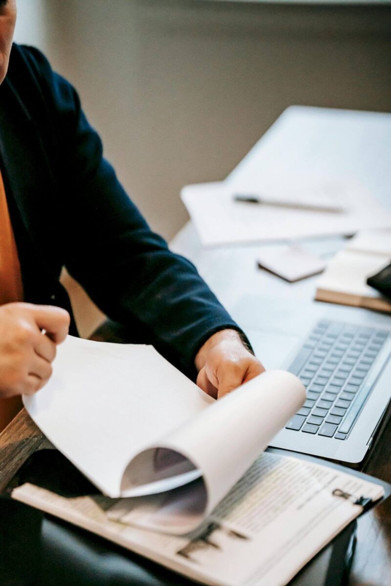 Person flipping through documents at a desk with a laptop, notebooks, and papers scattered around.