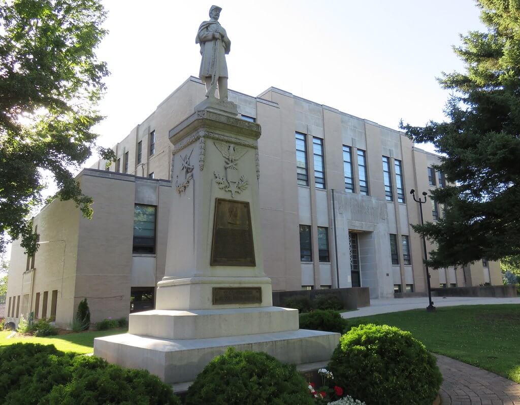 A stone statue of a soldier stands on a pedestal in front of a large building with multiple windows, surrounded by trees and bushes.