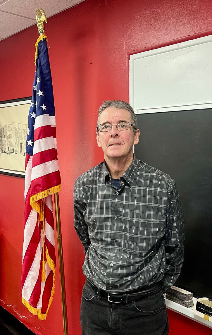 Man in a checked shirt stands beside an American flag and a blackboard in a room with red walls.