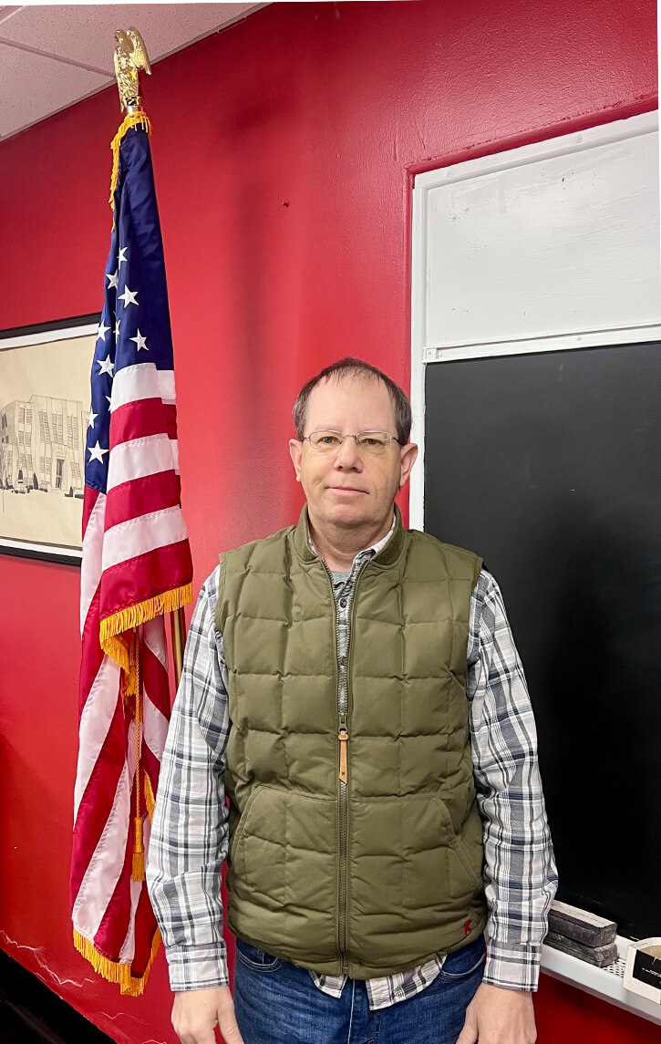 Man in plaid shirt and green vest stands in front of an American flag and blackboard, with a red wall background.