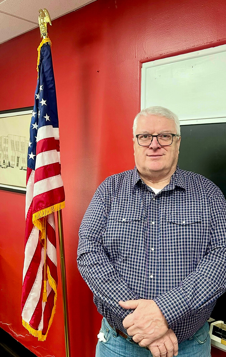 Man standing beside an American flag against a red wall.