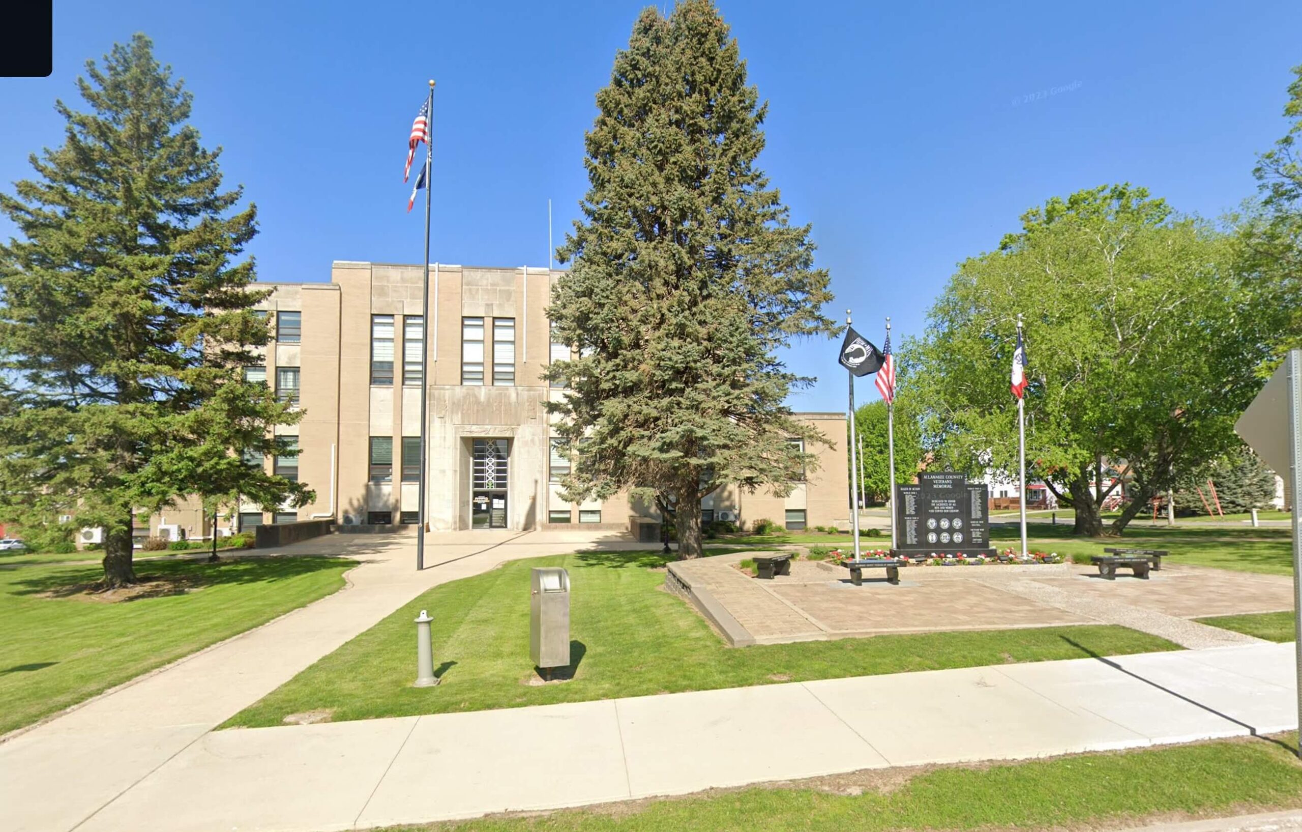 A stone building with flags and a memorial in front, surrounded by green grass and trees, under a clear blue sky.